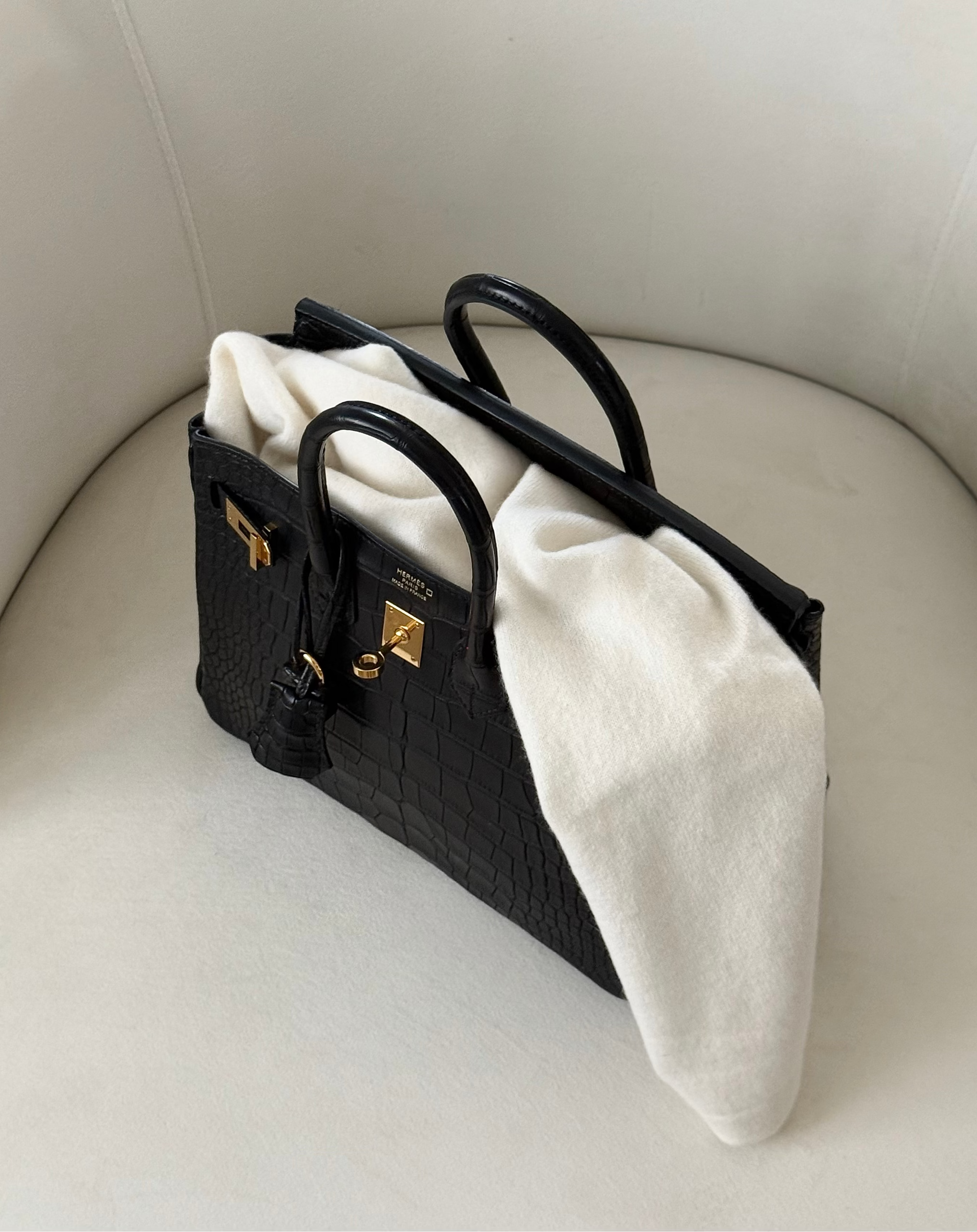 A black crocodile-embossed top-handle bag with gold hardware on a cream boucle chair, ivory knitwear draped beside it